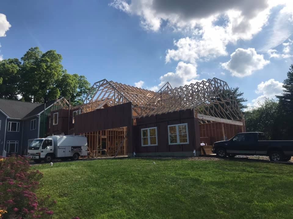 House under construction with exposed roof framing, trucks in front, and a grassy yard on a sunny day