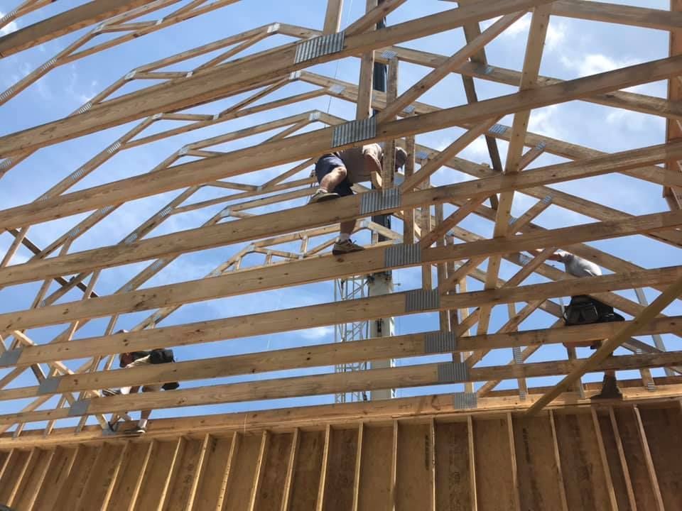 Workers on a wooden roof truss under construction, with one person kneeling on the framing.