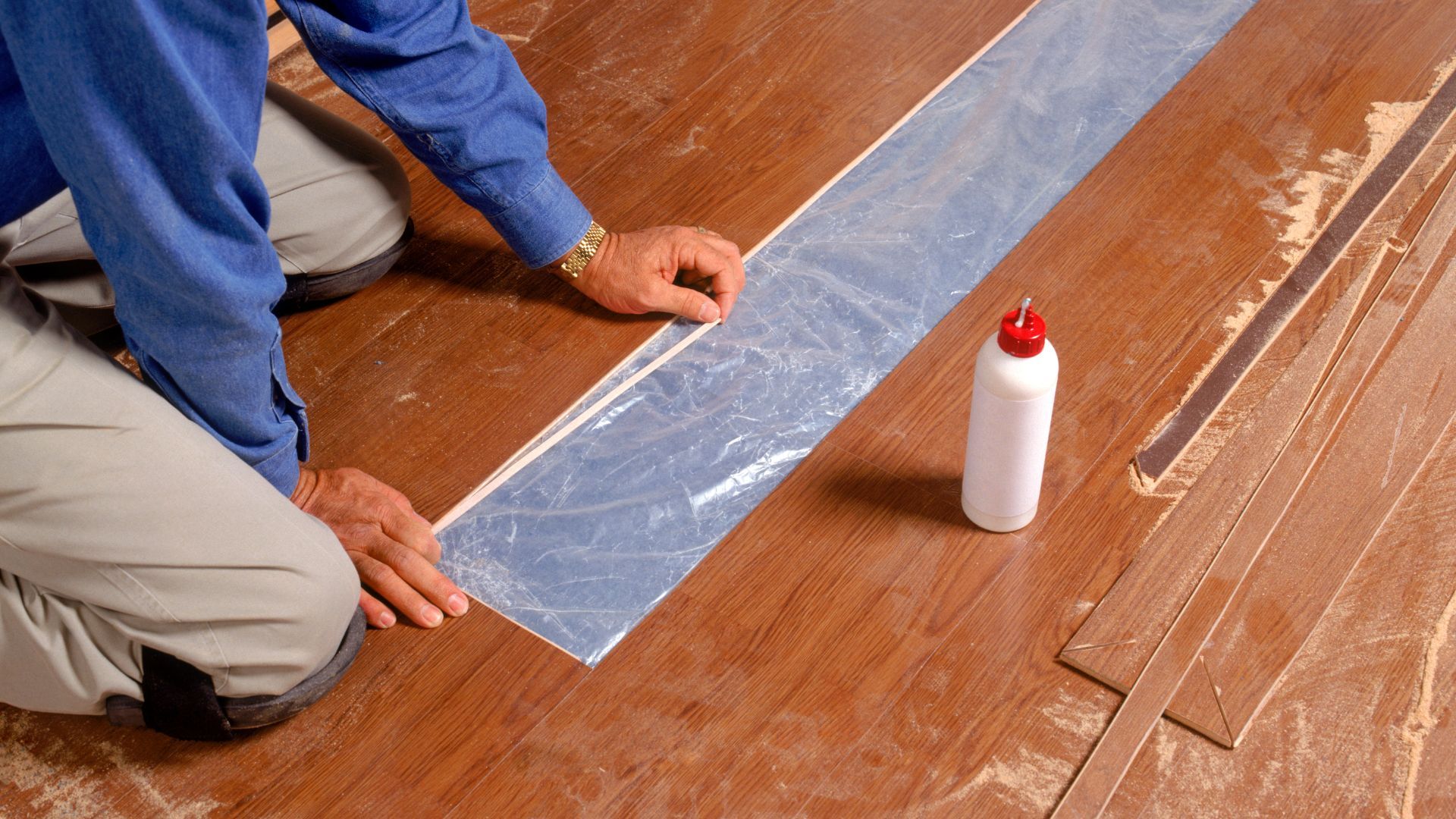 A man is kneeling down on a wooden floor next to a bottle of glue.