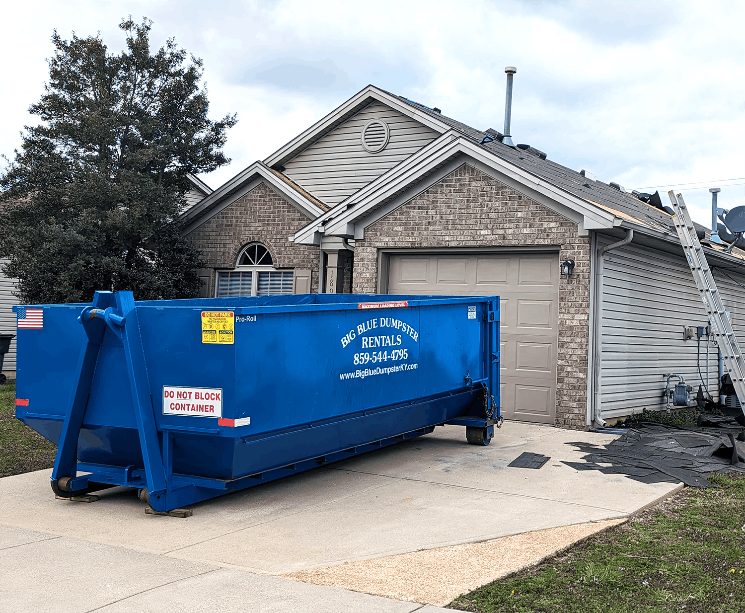 Blue dumpster in a suburban driveway in front of a brick house with a garage.