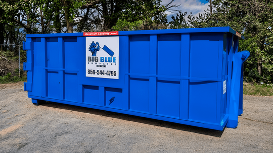 Blue dumpster with logo, parked on a gravel lot beside trees.