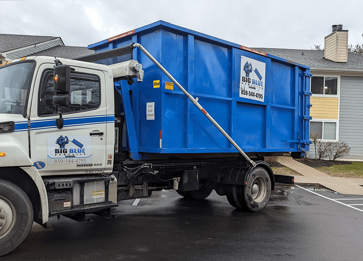 Blue dumpster truck parked on a wet street beside houses on a cloudy day