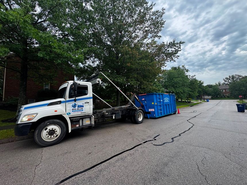 Tow truck pulling a blue dumpster along a cracked residential street under cloudy skies
