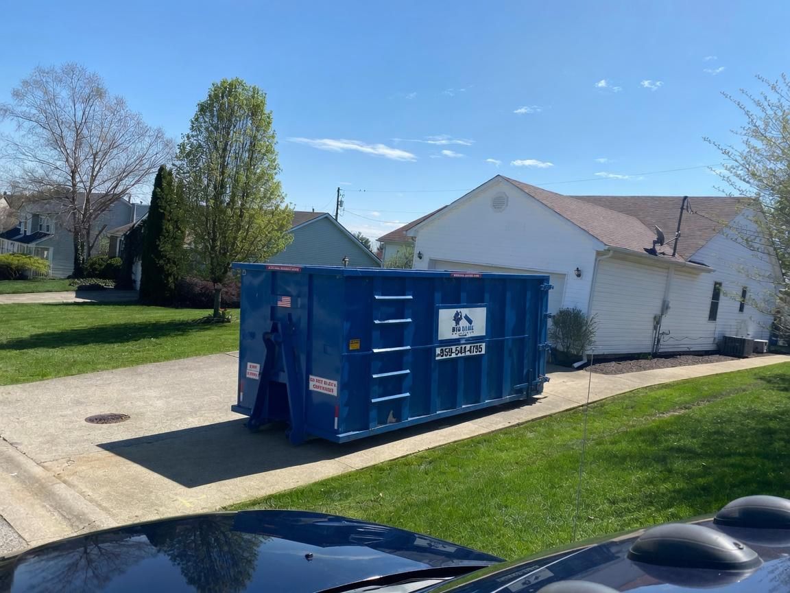 Blue dumpster on a driveway in a suburban neighborhood, with houses and trees in the background