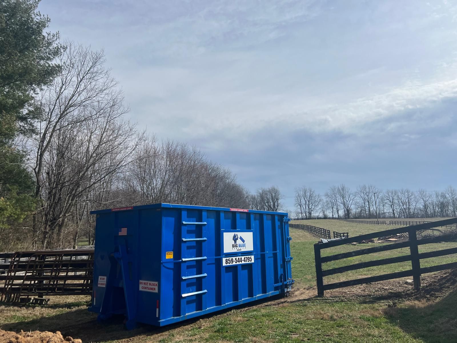 Blue dumpster beside an open wooden gate in a rural field under a cloudy sky