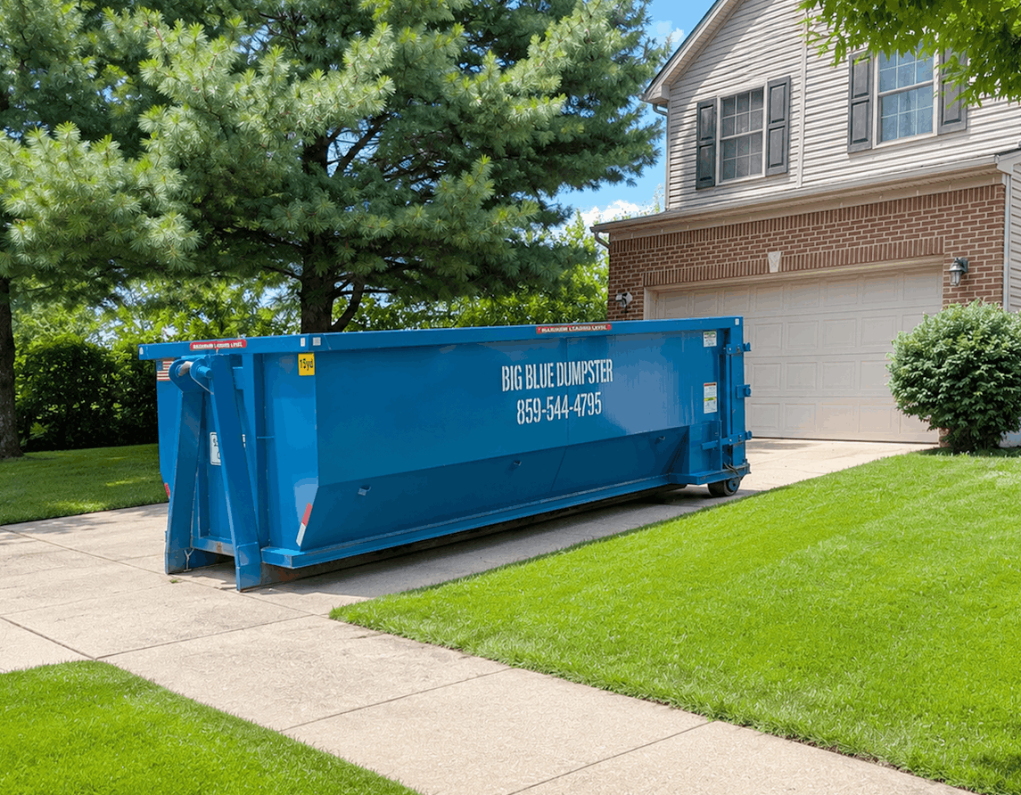 Blue dumpster on a driveway beside a suburban house and green lawn.