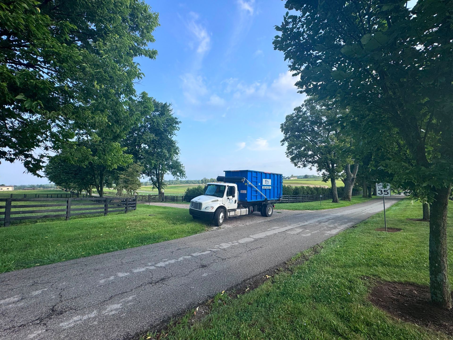 White truck with blue cargo bed parked on a country road beside green fields and trees under a blue sky