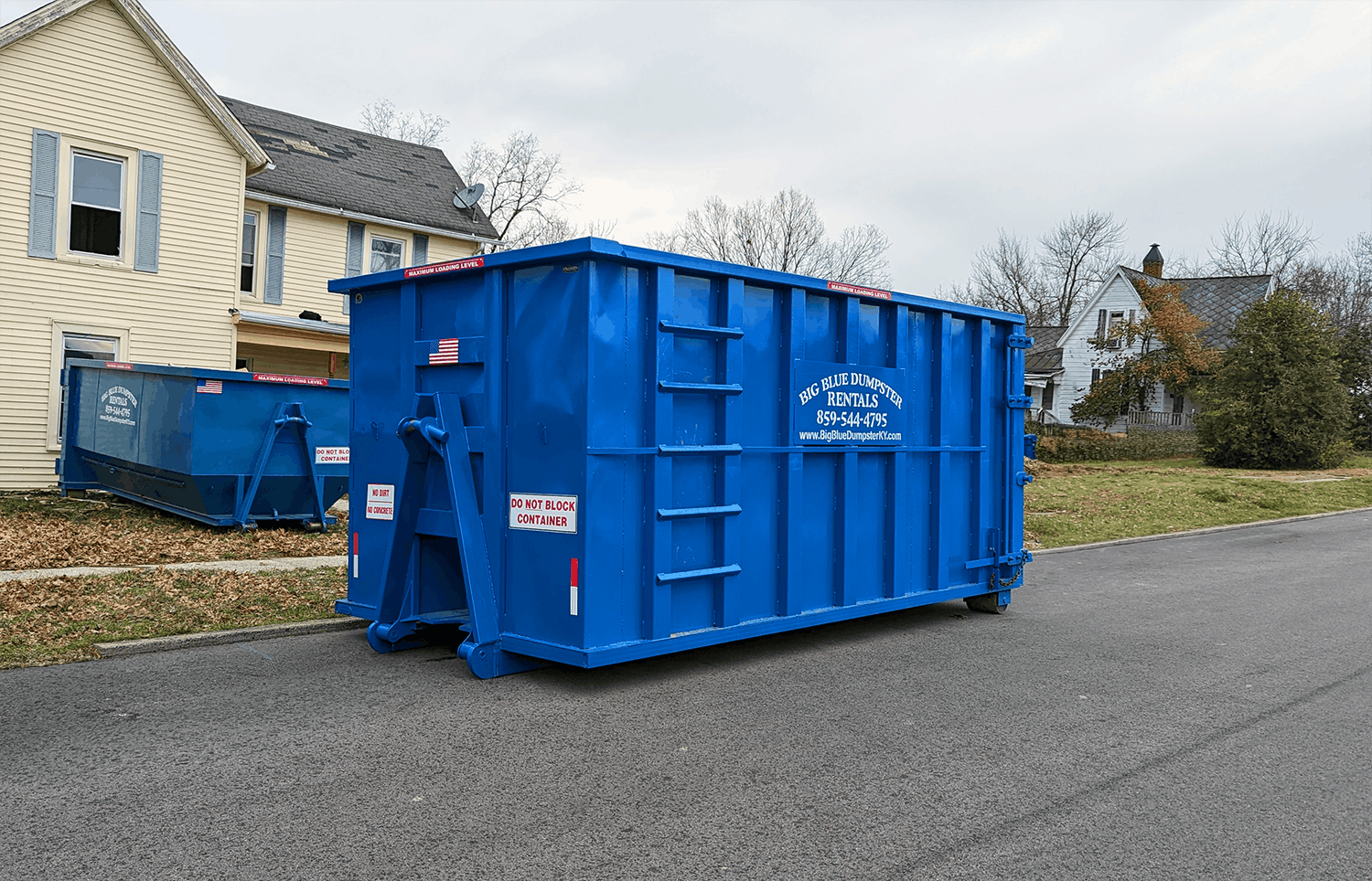 Blue dumpster bin on a residential street beside a house and another dumpster.