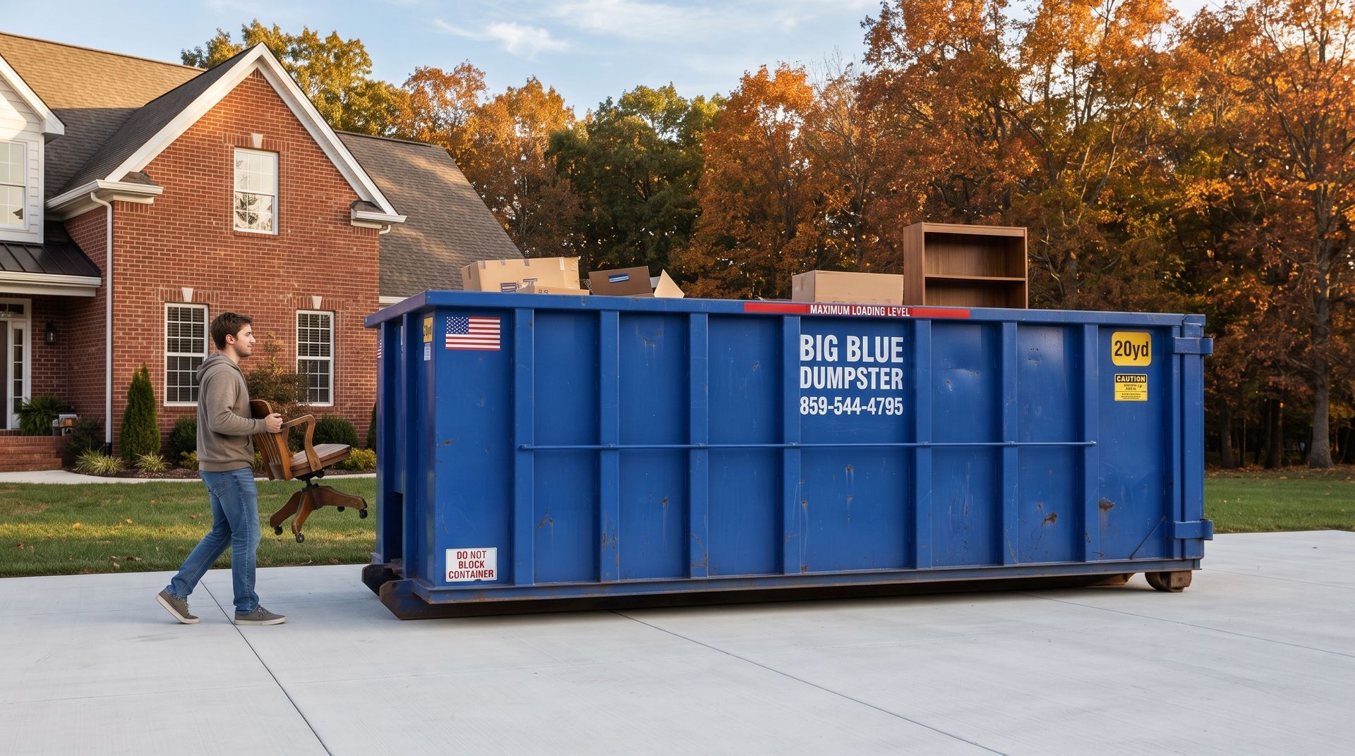 Worker moving a large blue dumpster on a suburban driveway with brick houses in the background