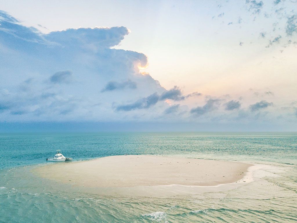 Sandy tropical sandbar in turquoise water under a pastel cloudy sky, with a small boat nearby