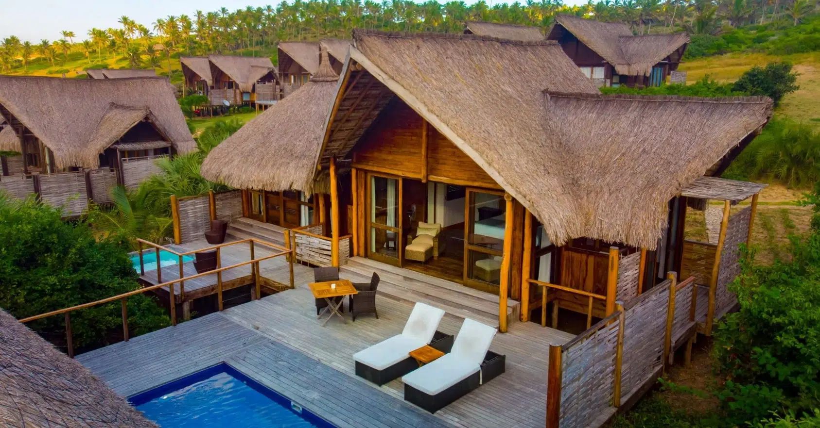 Luxury wooden bungalows with thatched roofs and private pools in a lush, tropical landscape.