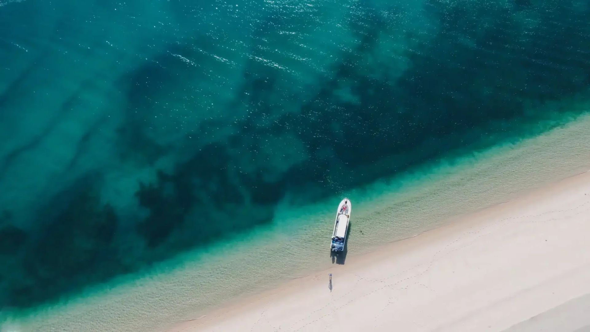 A small boat rests on the edge of a sandy beach next to turquoise ocean waters, viewed from directly above.