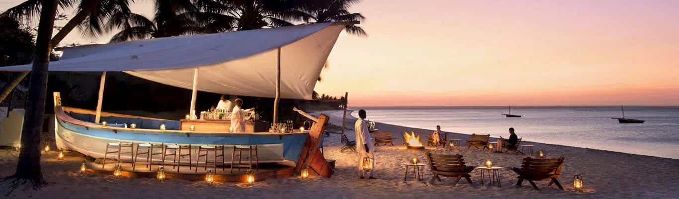 A blue boat converted into a beach bar, sheltered by a white canopy, sits on sandy ground at sunset near the ocean.
