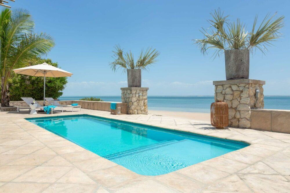 A wooden pergola with lounge seating overlooks a turquoise infinity pool and a sandy beach under a clear blue sky.