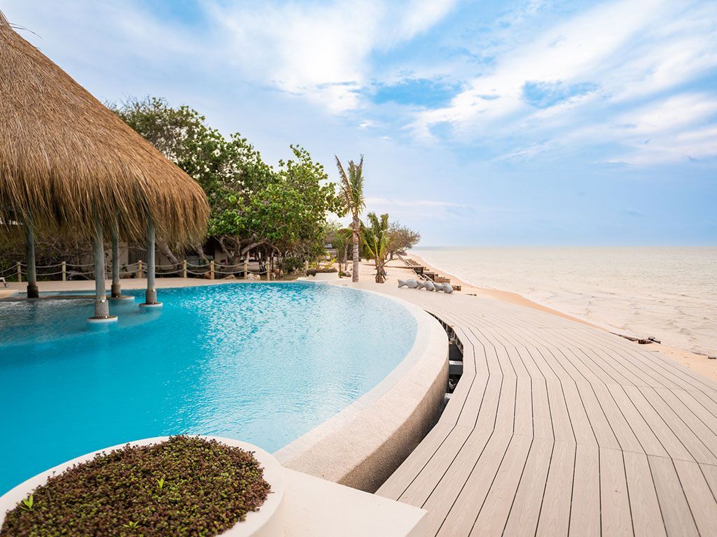 Resort pool beside a sandy beach with thatched shade and turquoise water under a bright sky