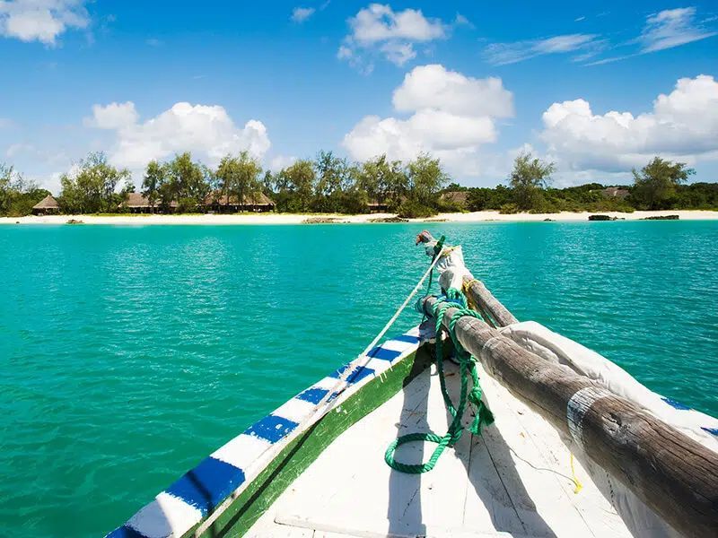 The bow of a boat with blue and white stripes approaches a sandy beach with palm trees under a bright blue, cloudy sky.