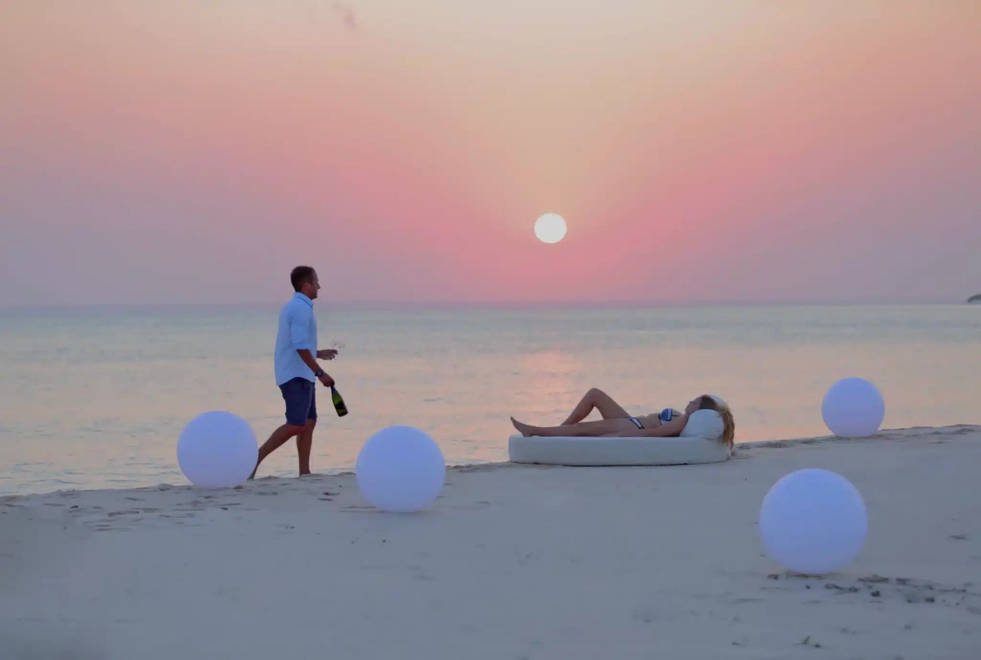A person walks toward another lying on a beach lounger at sunset, surrounded by glowing white orbs on the sand.