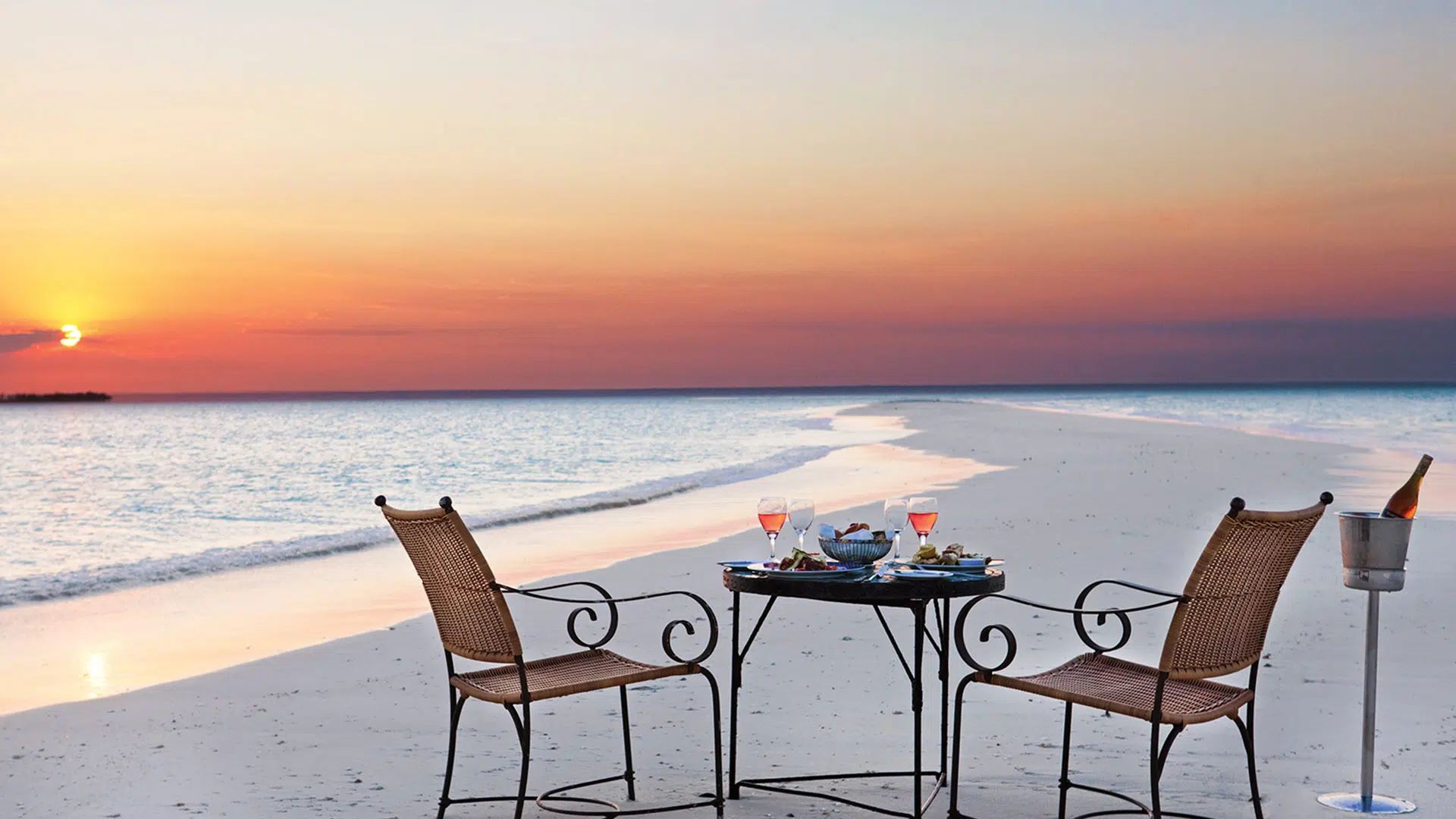 A table for two with champagne on a white sand beach at sunset, with the ocean stretching to the horizon.