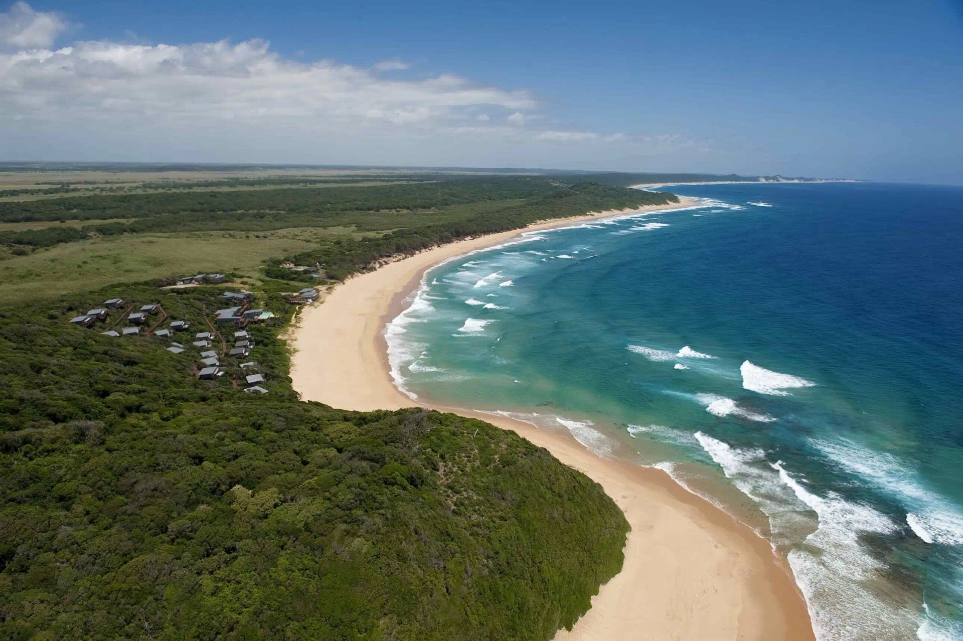 An aerial view of a tropical sandy beach bordering a dense green forest, with a small resort nestled in the trees.