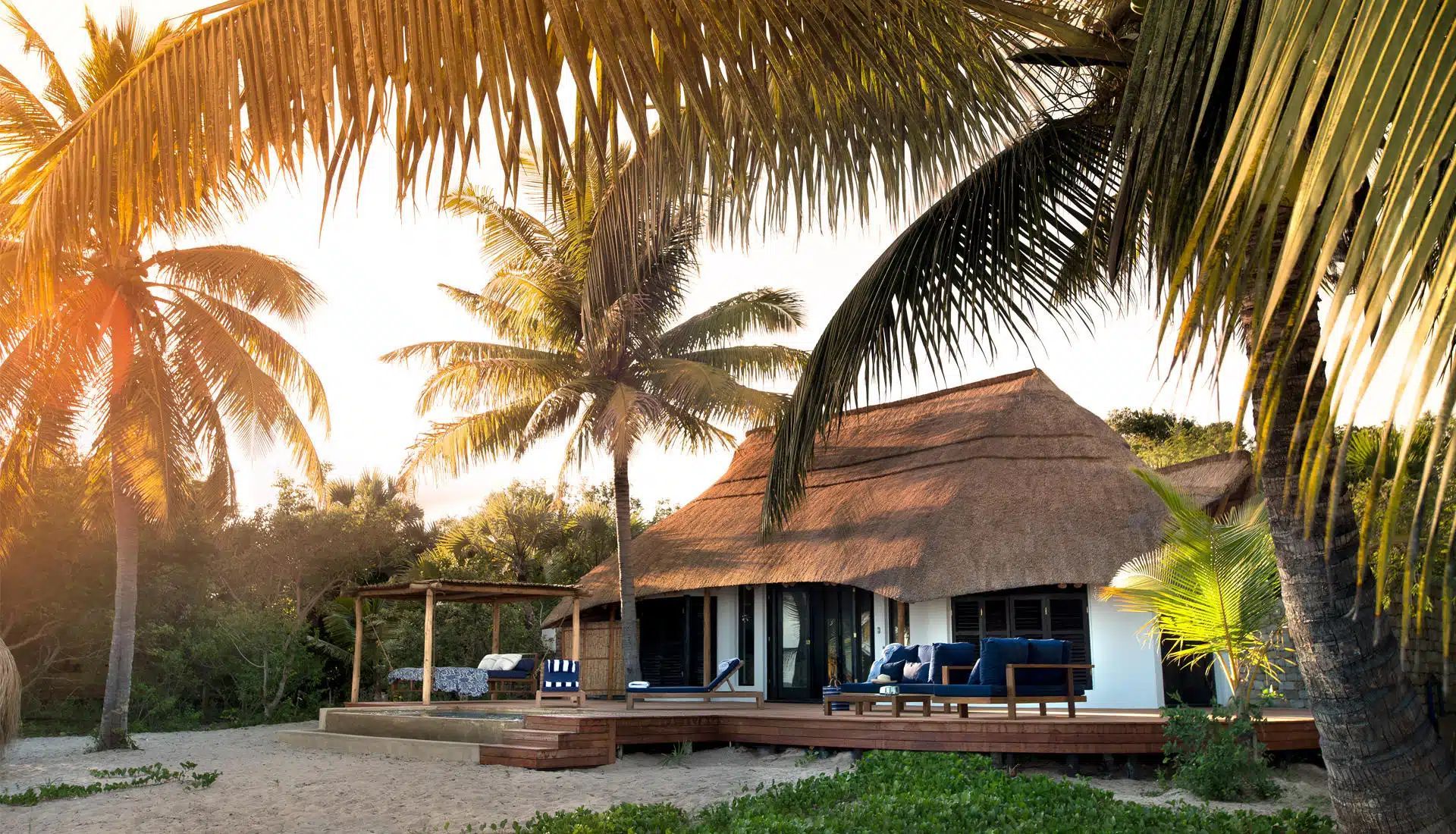 A thatched-roof bungalow on a tropical beach surrounded by palm trees during a golden sunset.
