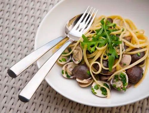 A bowl of linguine with clams, garnished with fresh greens, set on a woven surface with a fork and spoon.