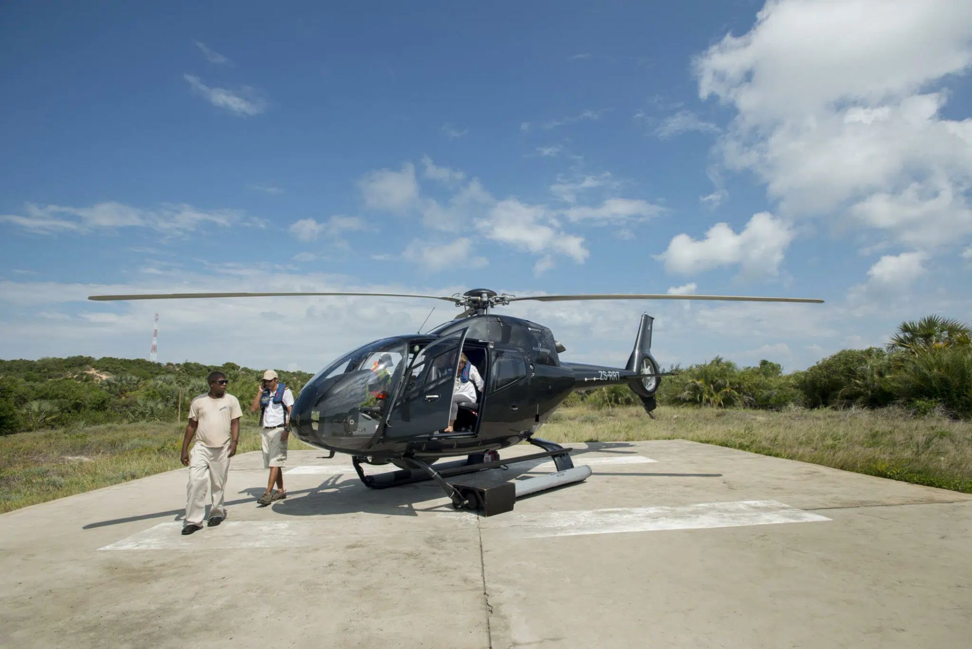 Two people walk toward a small, dark helicopter parked on a concrete helipad under a bright, sunny blue sky.