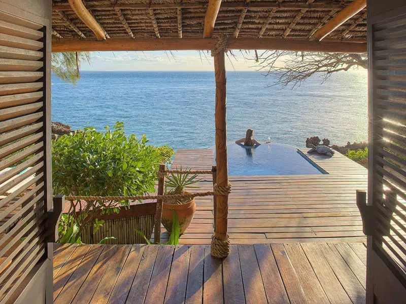 A person relaxes in an infinity pool overlooking the ocean from a wooden deck under a thatched roof.