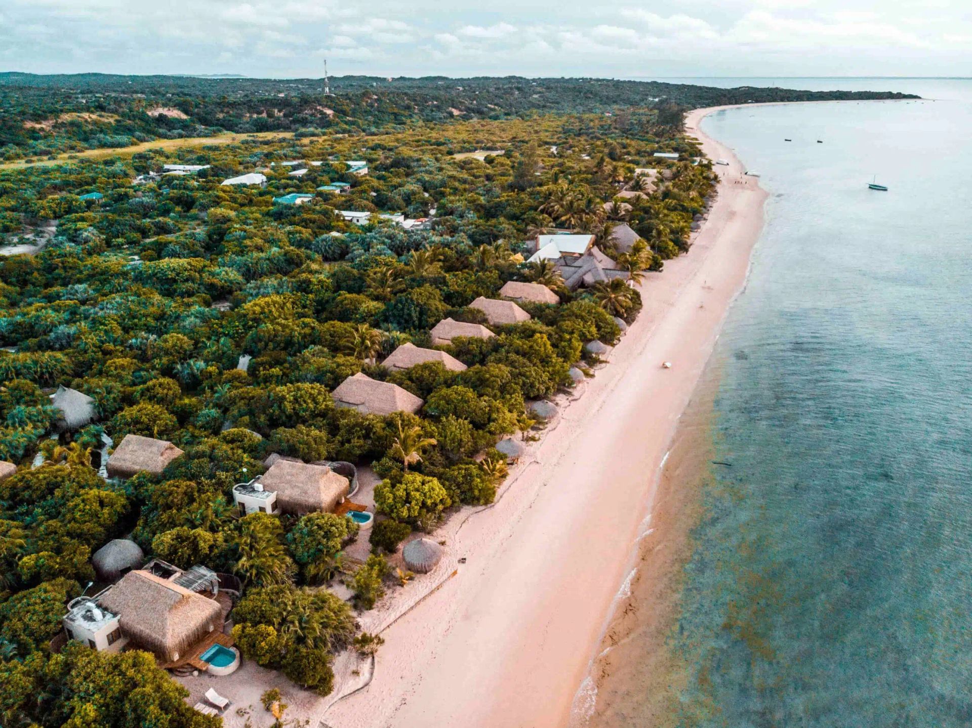 Aerial view of a sandy beach lined with thatched-roof villas tucked into a lush green forest along a calm, blue ocean.