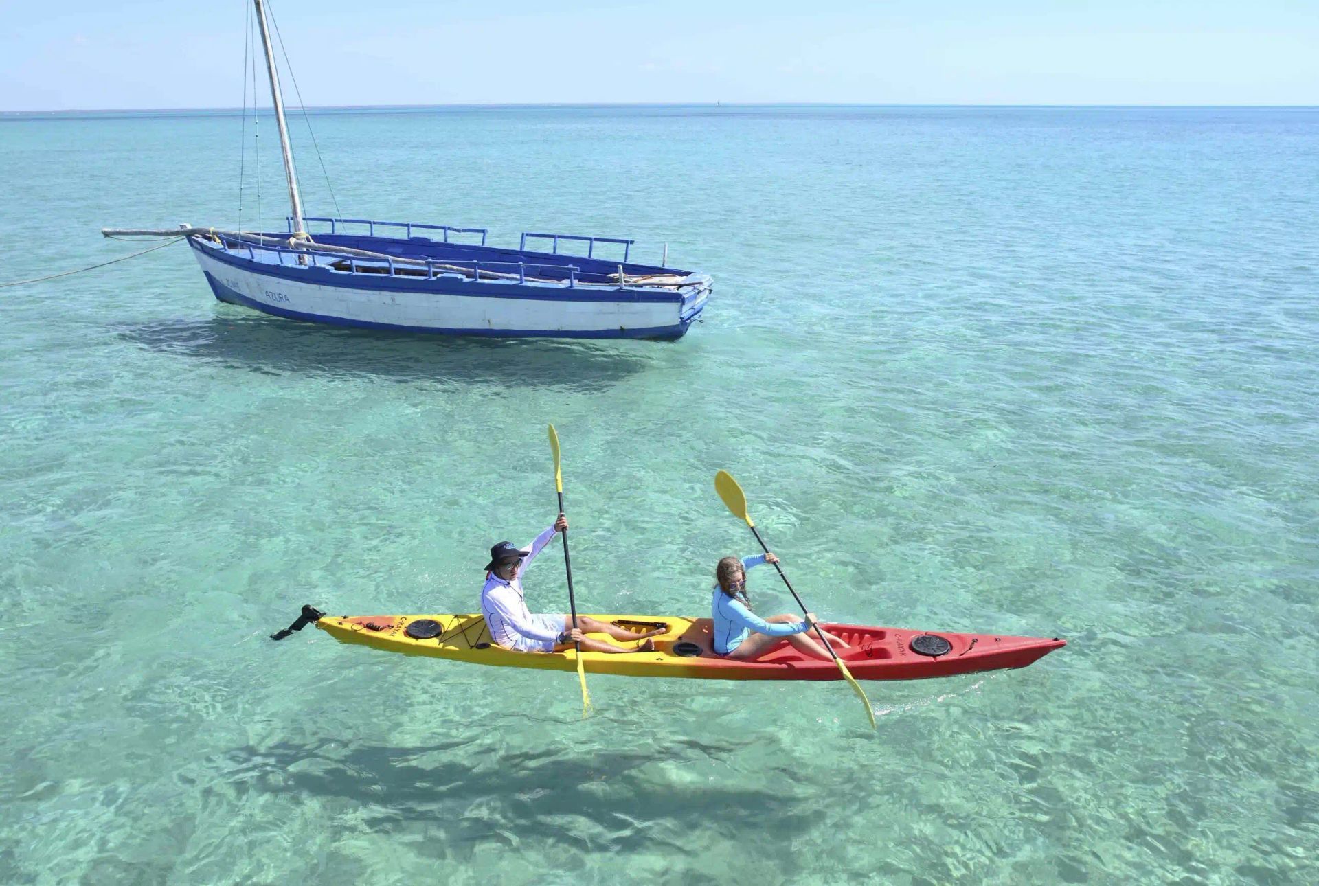 Two people paddle a red and yellow kayak in clear, calm ocean water near a small blue and white sailboat.