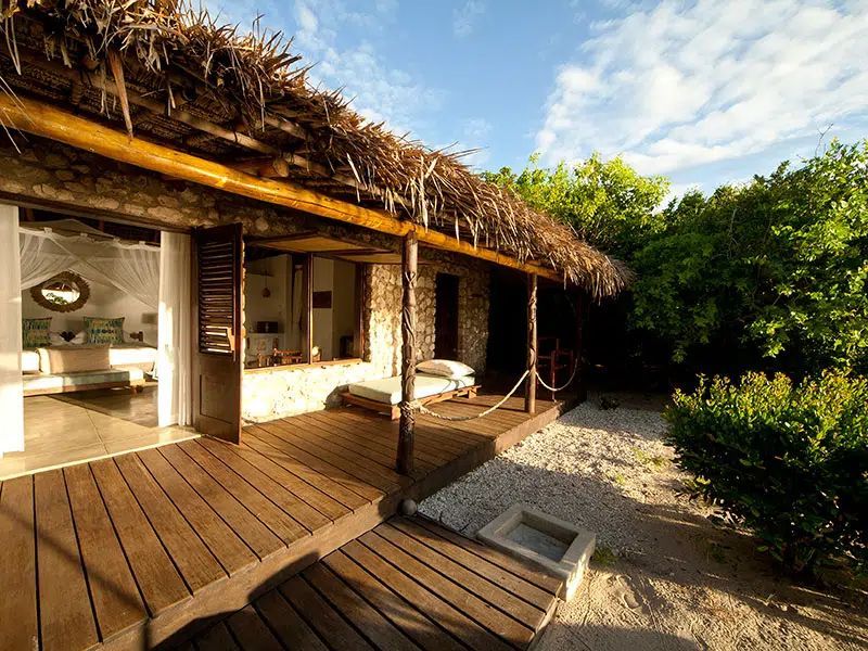 A rustic villa with a thatched roof, wooden deck, and open doorway revealing a bright interior, set near lush greenery.