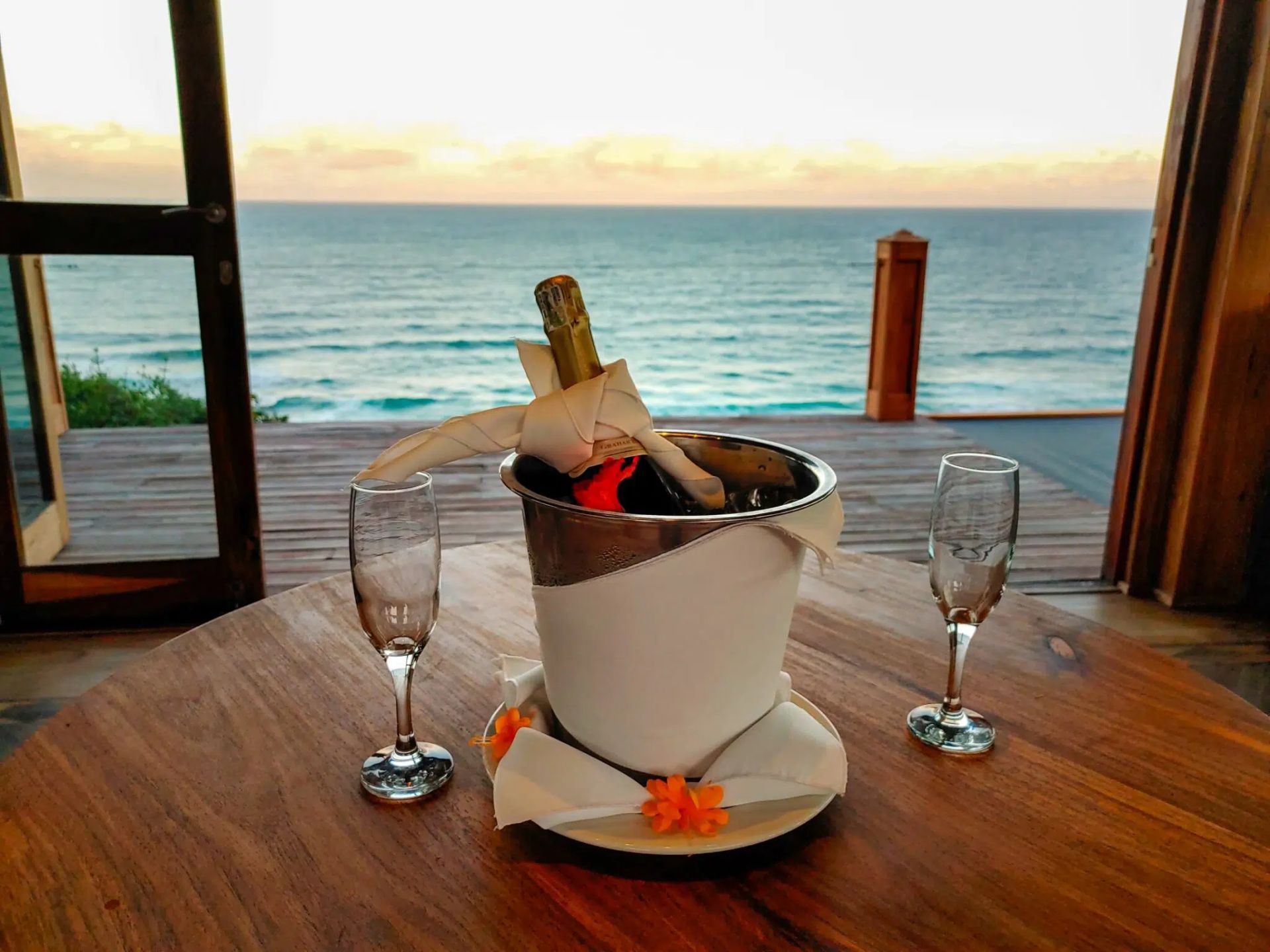 A bottle of champagne in an ice bucket with two flutes on a wooden table, overlooking the ocean at sunset.
