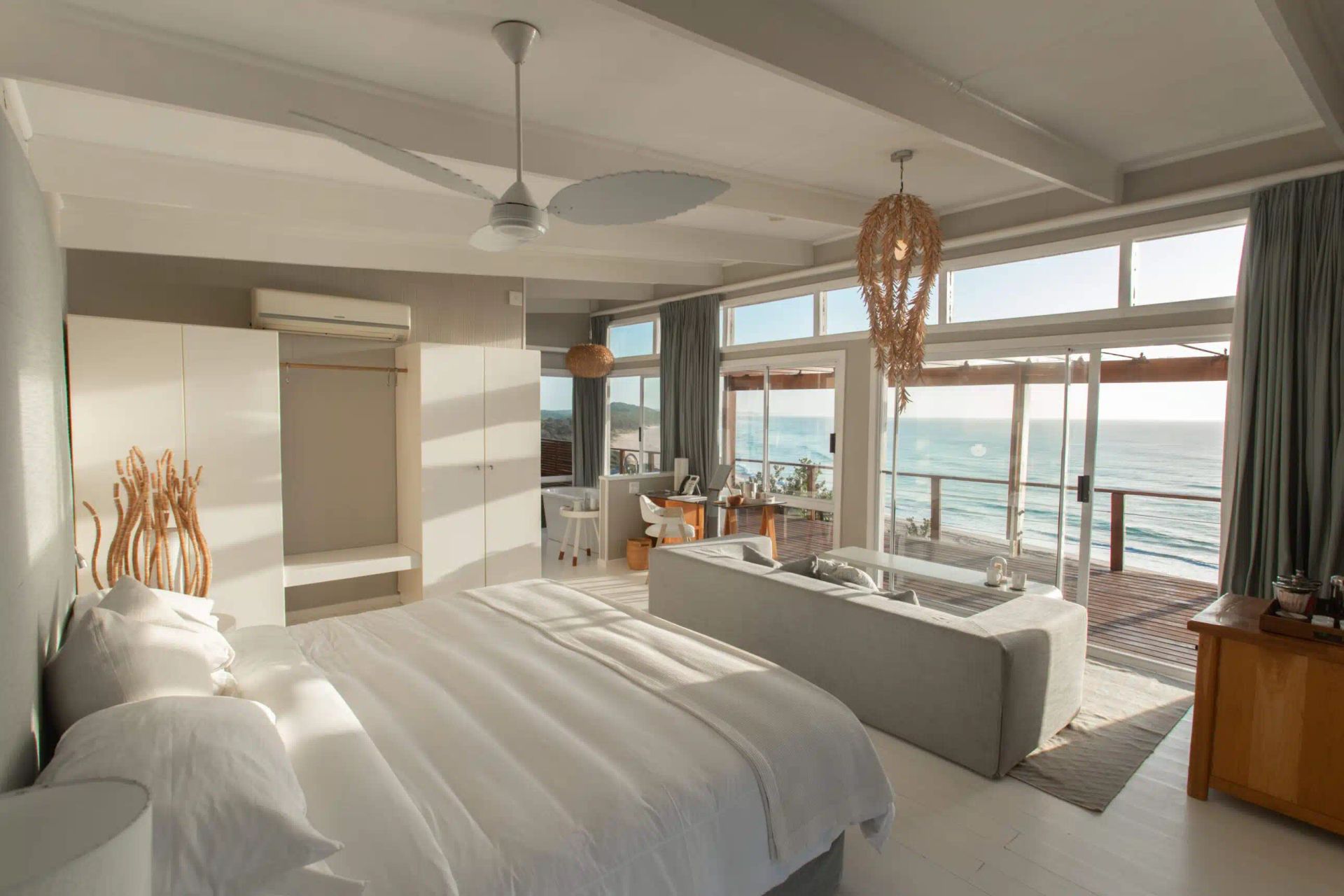 A bright, minimalist bedroom with a white bed, a sofa facing a large window with ocean views, and a hanging light fixture.