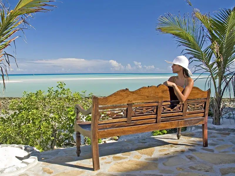 A person wearing a sun hat sits on a wooden bench facing a calm, turquoise tropical sea under a clear blue sky.