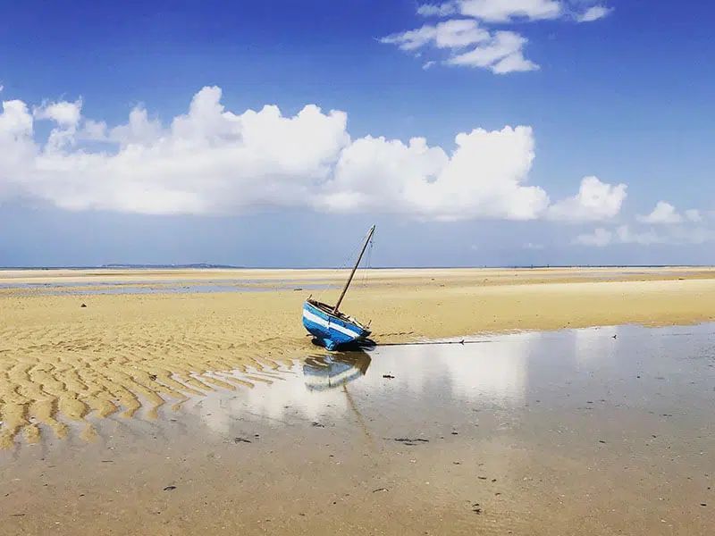 A small blue boat rests on a vast, sandy beach at low tide under a bright blue, cloudy sky.