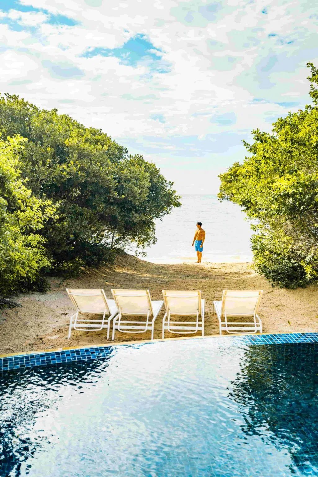 Four lounge chairs on a sandy path facing an infinity pool, with a person looking out toward the ocean at the horizon.