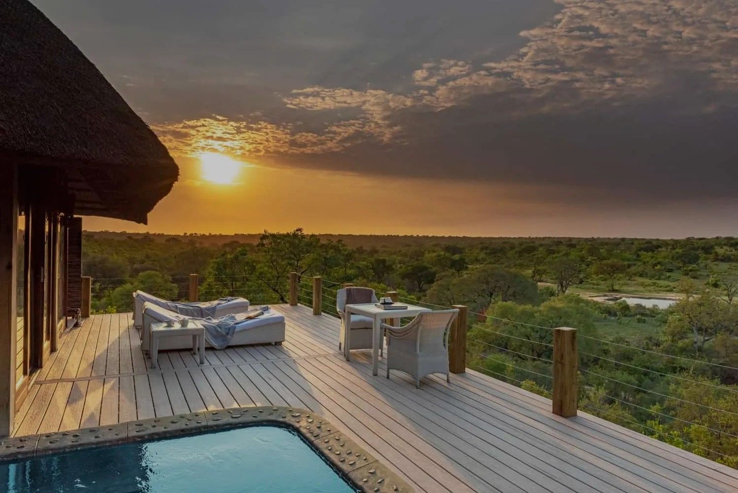 A sunset over a savanna landscape, viewed from a wooden lodge deck featuring lounge seating and a small pool.