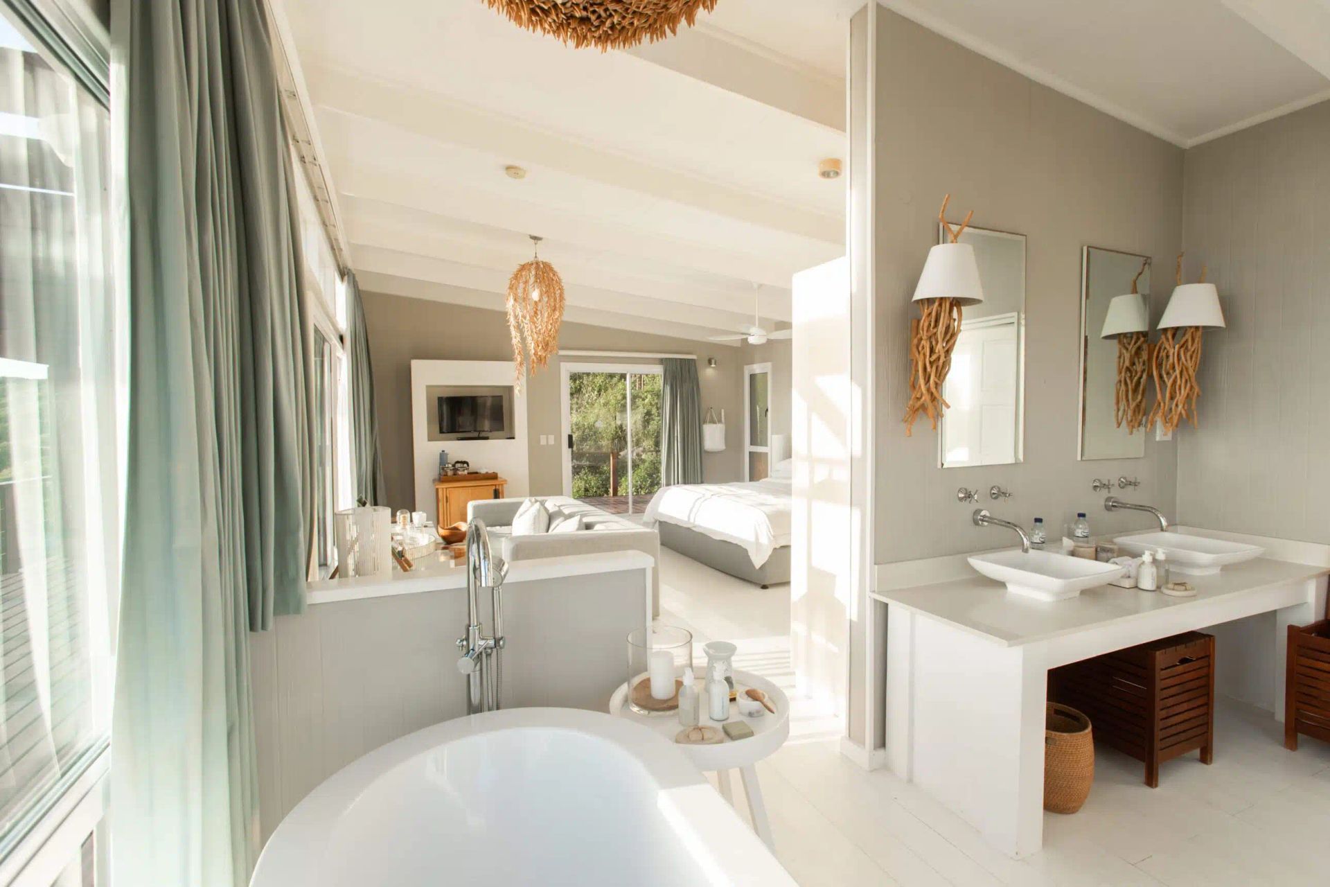 A modern, airy bathroom with a white freestanding tub, double vanity, and soft green curtains, opening into a bedroom.