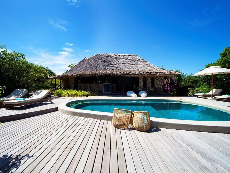 A swimming pool with two wicker baskets on the edge, set on a wooden deck by a thatch-roofed villa under a clear blue sky.
