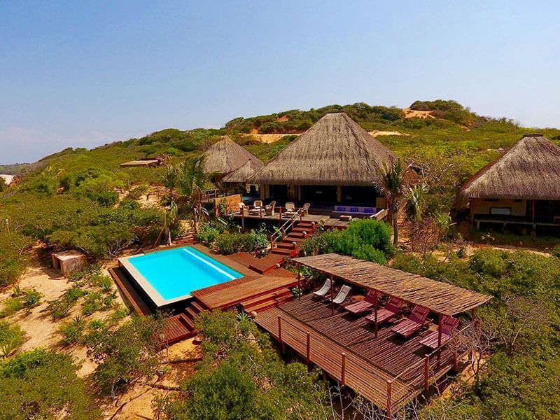 A luxury resort with thatched-roof villas, a bright blue swimming pool, and a shaded wooden deck with lounge chairs.