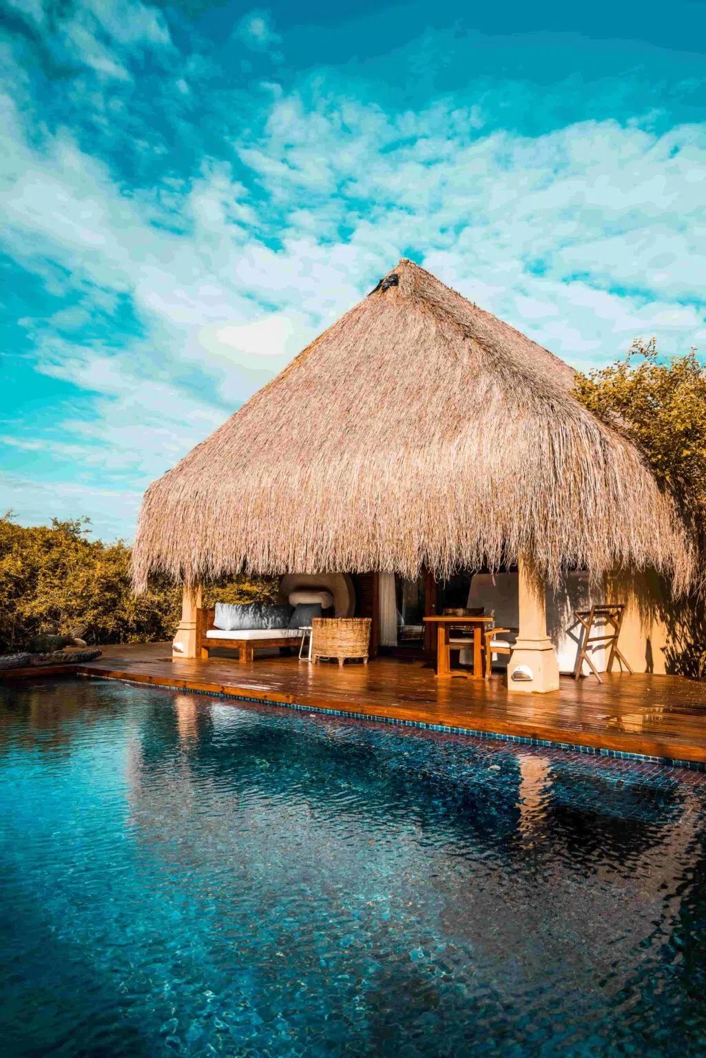 A thatched-roof hut with a patio and private pool under a vibrant blue sky with fluffy white clouds.