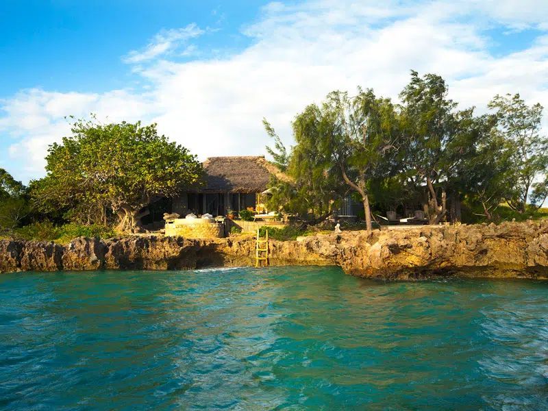 A tropical home with a thatched roof sits atop a jagged rock coastline overlooking turquoise water under a blue sky.