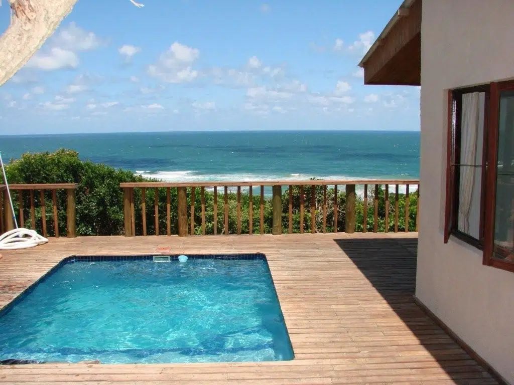 A swimming pool on a wooden deck overlooking a beach and the blue ocean under a bright, sunny sky.
