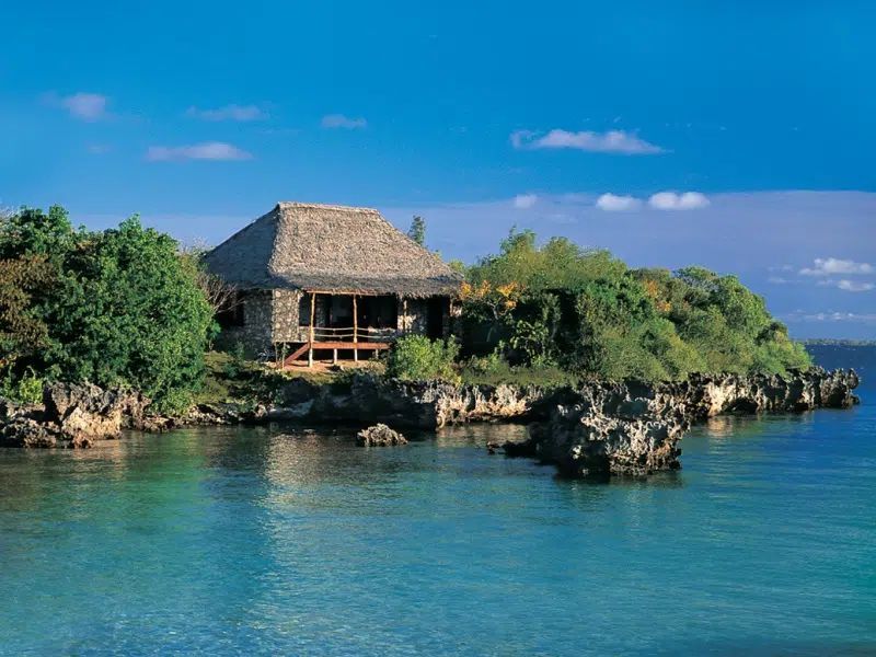 A thatched-roof cabin nestled in lush greenery on a rocky tropical island shore overlooking calm, turquoise water.