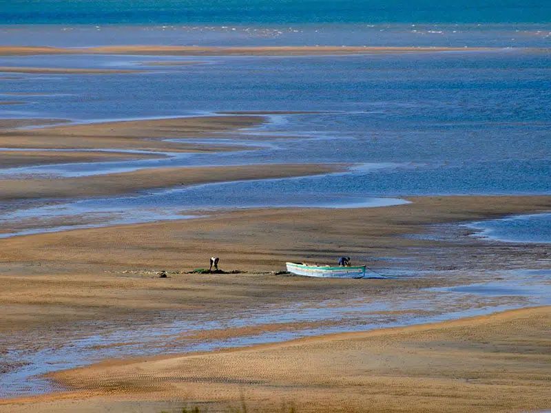 A small boat sits on expansive, sandy tidal flats under a clear blue sky, with a person working nearby.