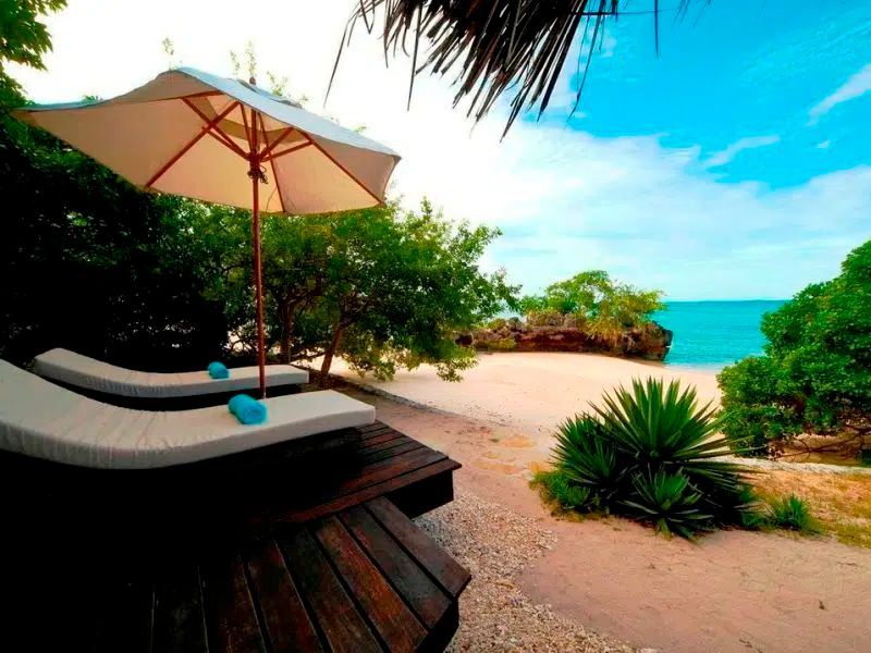 Two lounge chairs under an umbrella on a wooden deck overlooking a sandy beach and turquoise ocean.