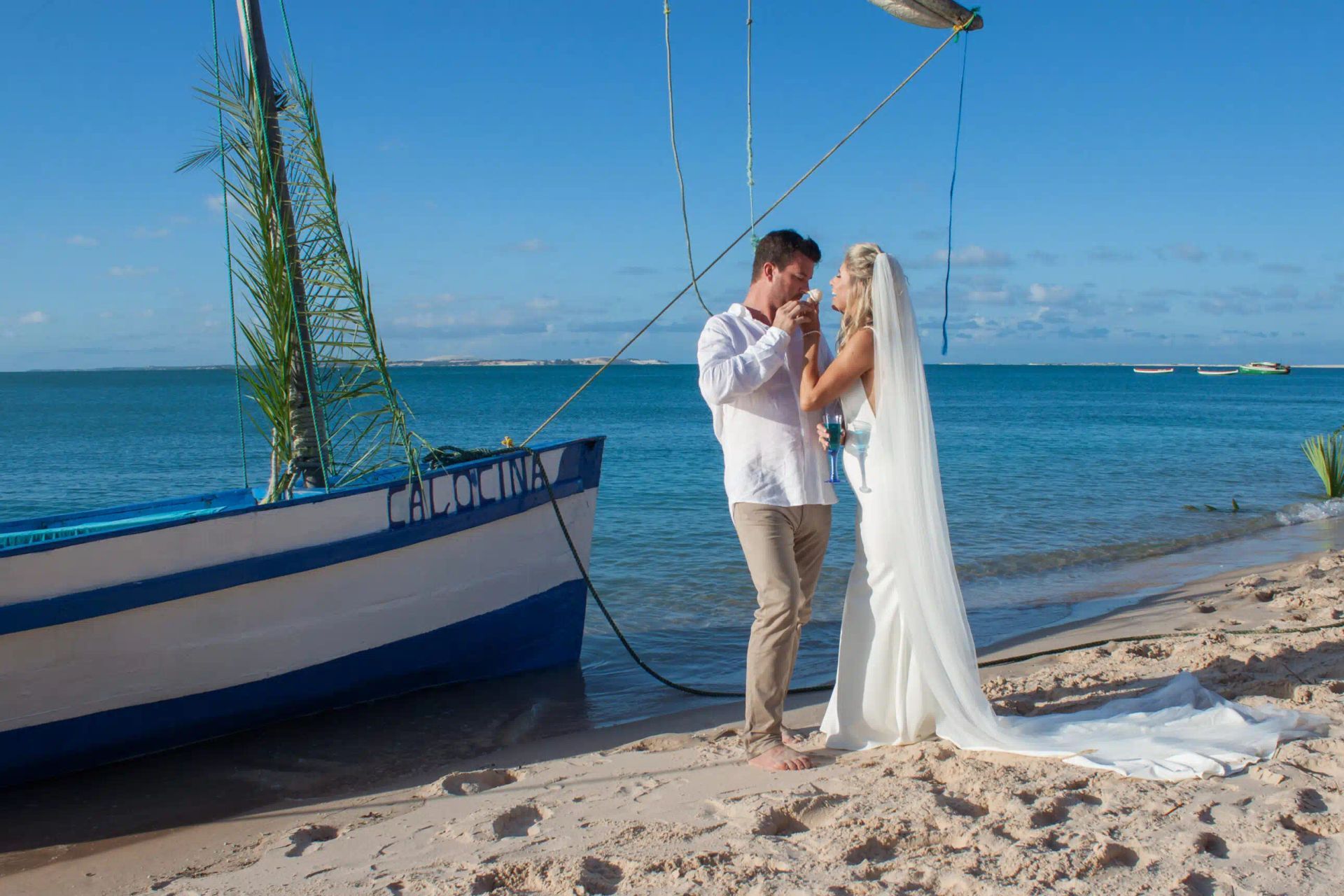 Bride and groom kissing on a sandy beach beside a blue-and-white boat and calm ocean