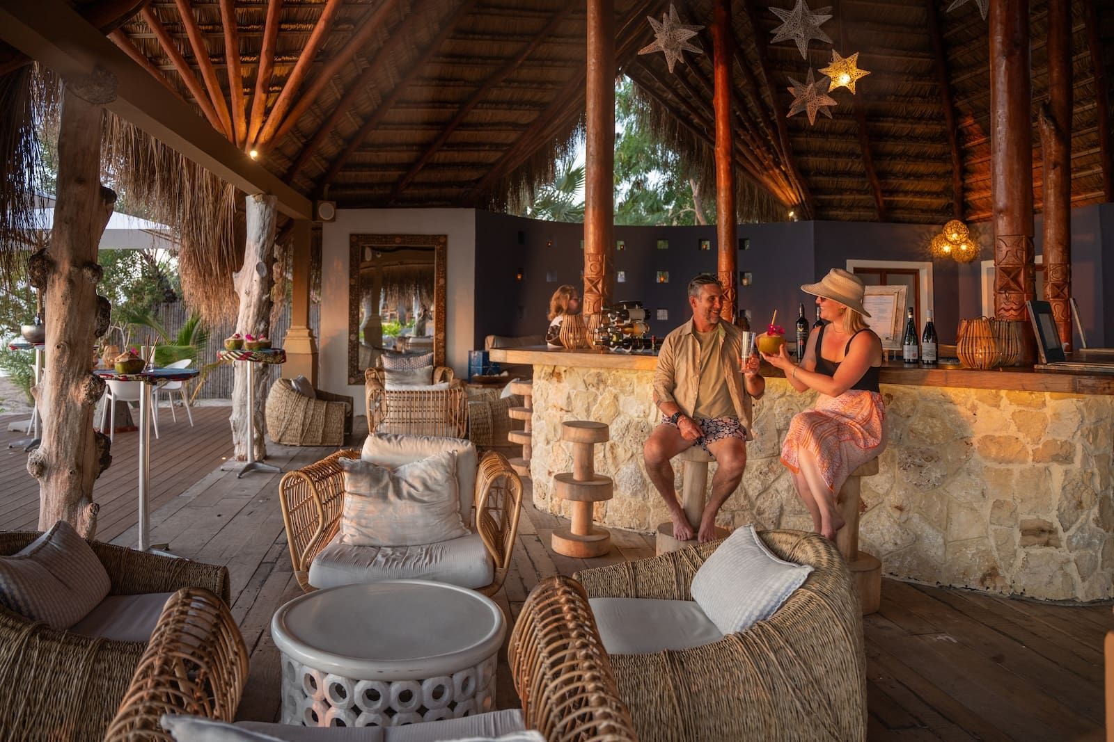 Open-air stone bar with wicker chairs and two people sitting in a tropical lounge at sunset