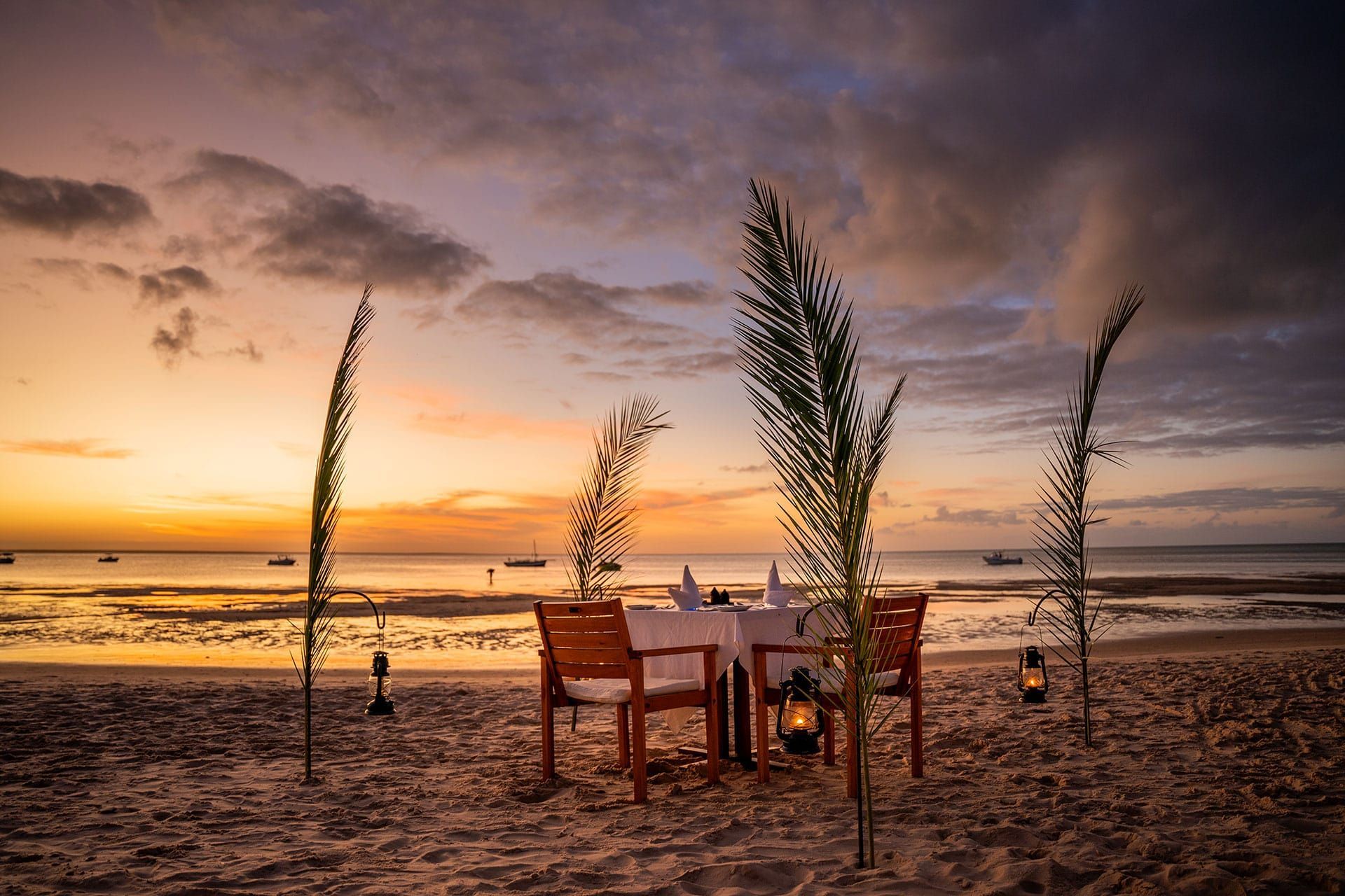 Beach dining table at sunset with lanterns and tall grasses beside the ocean