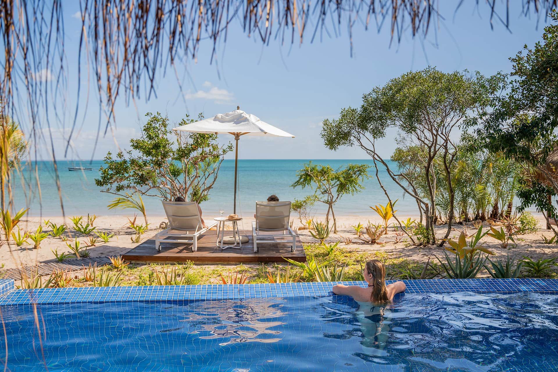 Infinity pool overlooking a tropical beach with lounge chairs, umbrella, and two people relaxing in the water