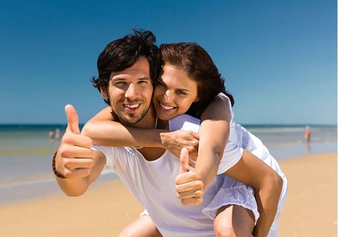 Smiling couple giving thumbs-up on a sunny beach, with blue sky and ocean in the background